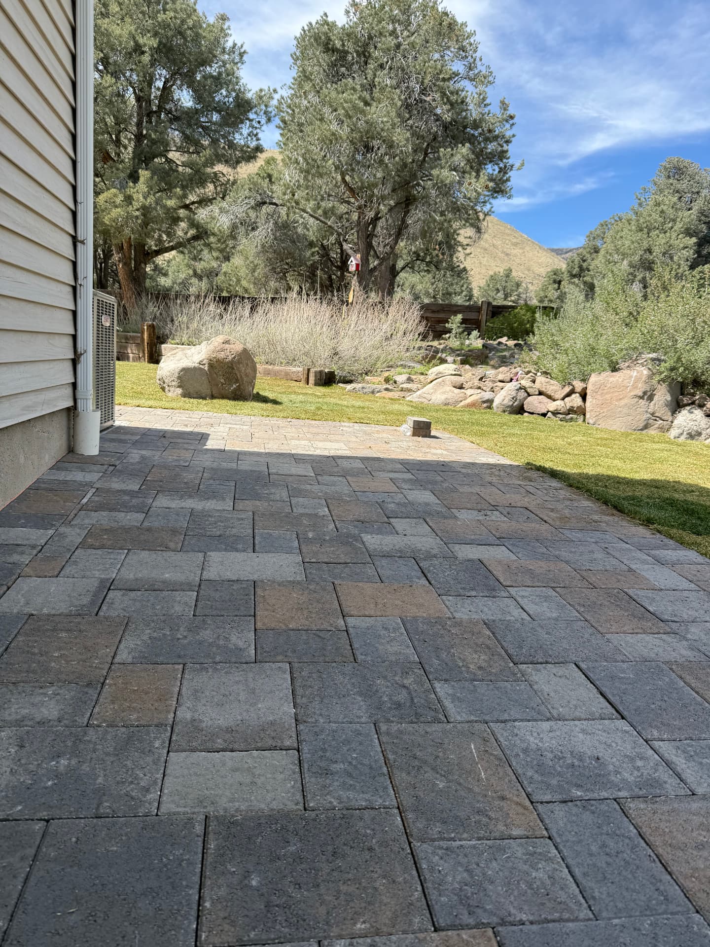 Wide paver patio along the house with lawn and boulders beyond